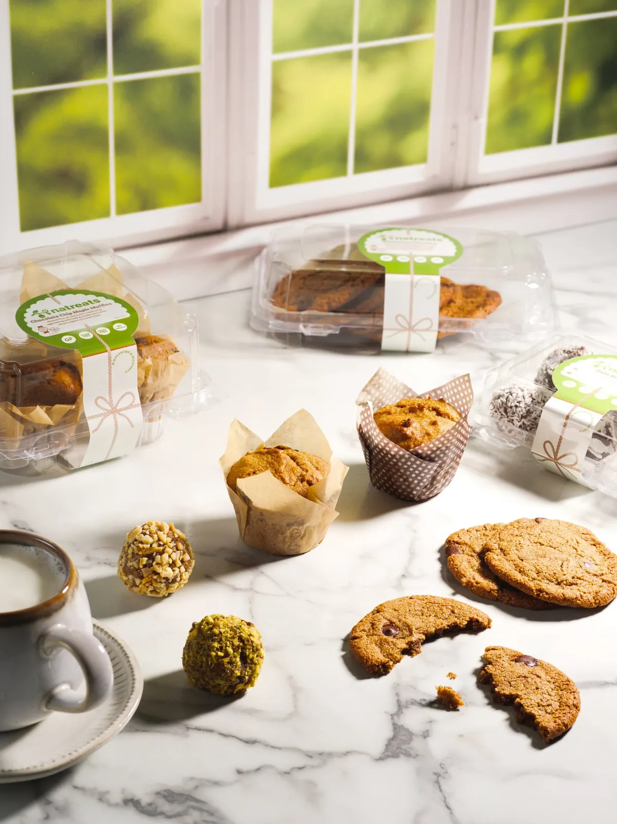 Natreats cookies, muffins and date balls arranged on a sunlit marble counter beside a warm cup of milk